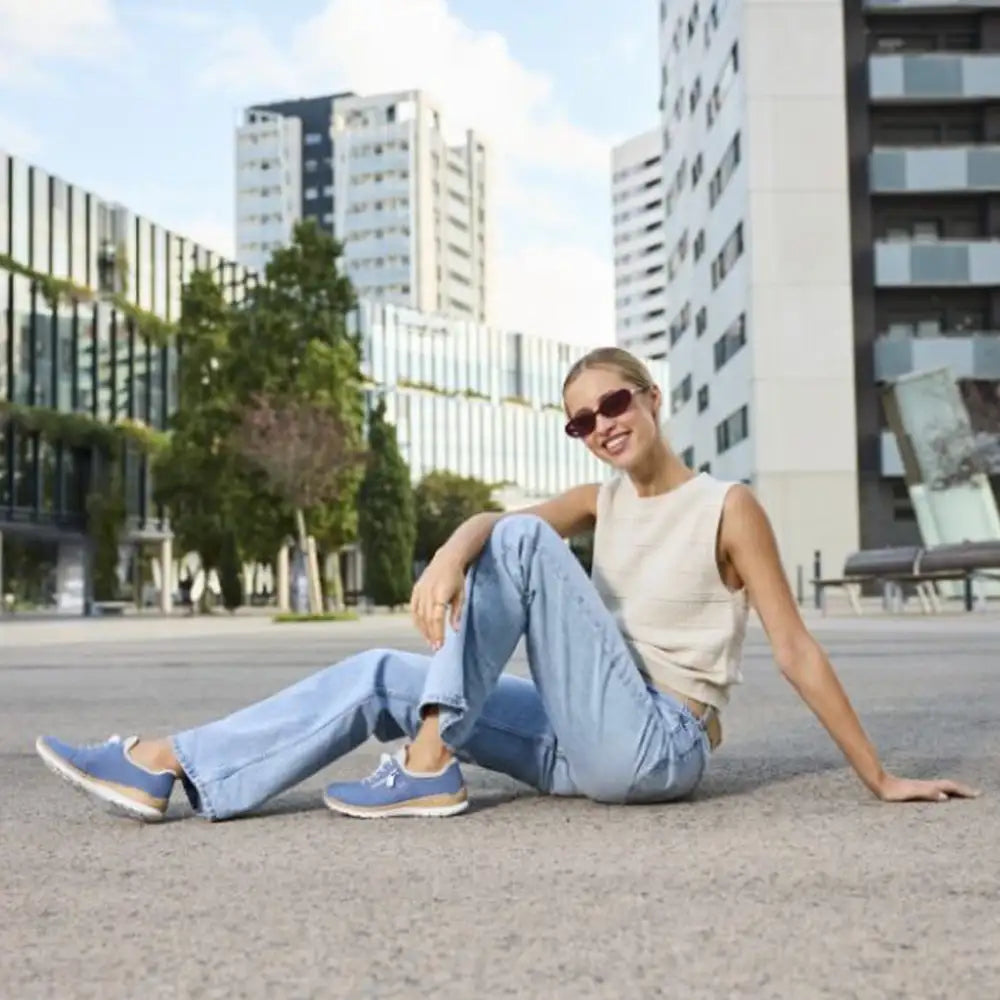Woman sitting on a city street wearing sunglasses and casual clothing with modern buildings in the background.